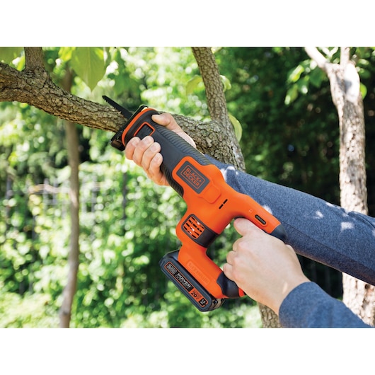 Scie sauteuse BLACKDECKER BDCR20C coupant une branche d’arbre en extérieur, outil électrique orange et noir.